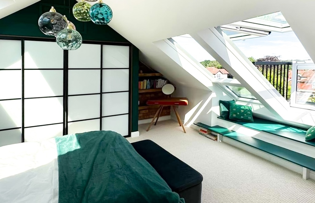 Modern attic bedroom with green and white decor, sloped ceiling, large skylight windows, built-in window seat, and pendant lighting.