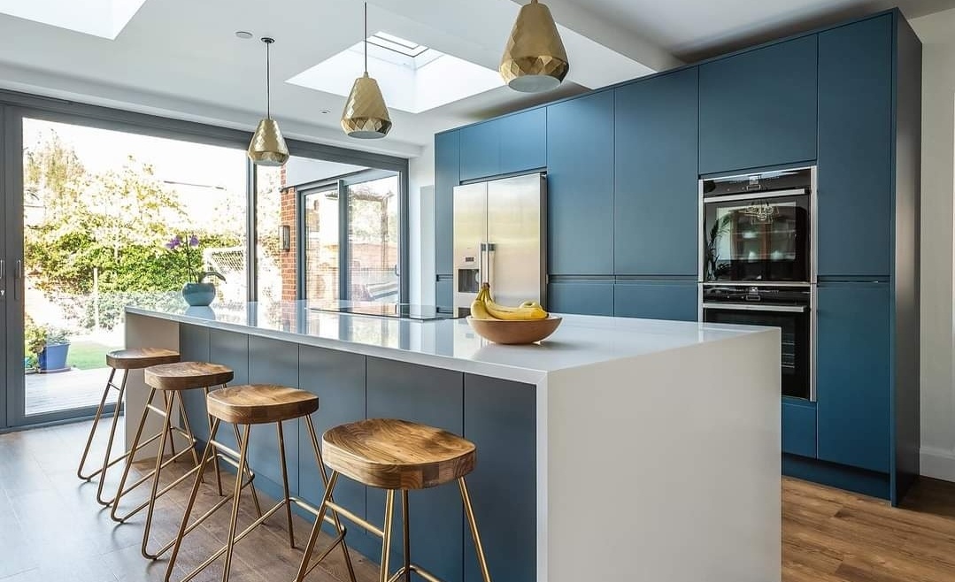 Contemporary kitchen with navy cabinets, white island, wooden bar stools with gold legs, skylights, and garden-facing glass doors.