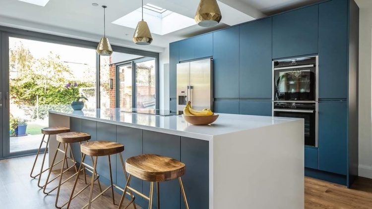 Contemporary kitchen with navy cabinets, white island, wooden bar stools with gold legs, skylights, and garden-facing glass doors.