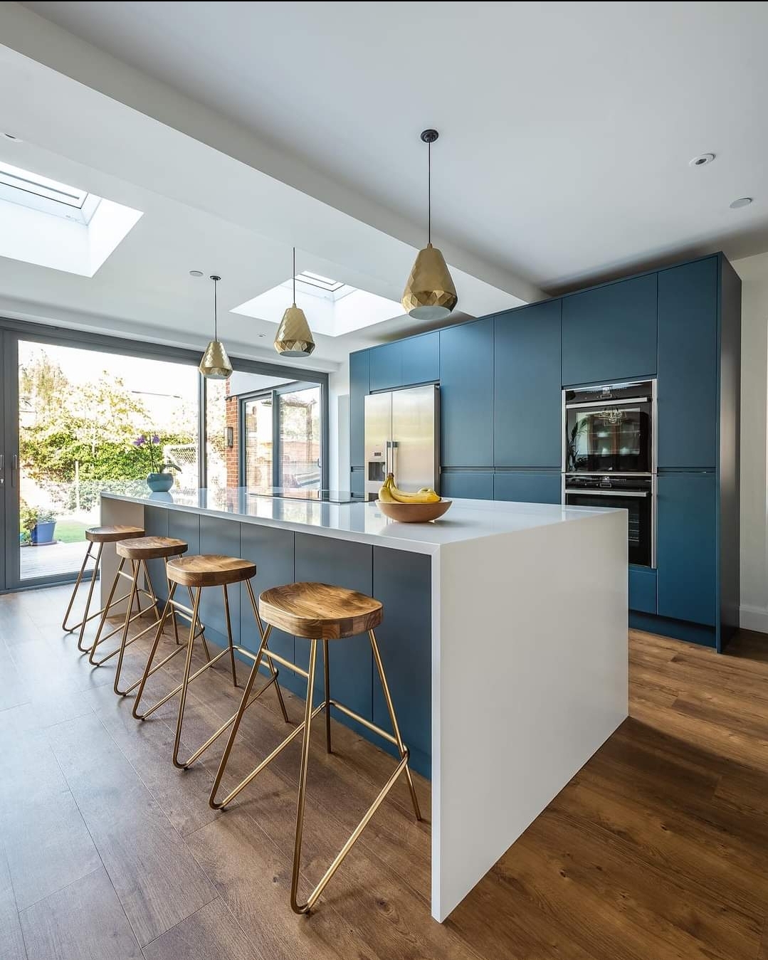 Contemporary kitchen with navy cabinets, white island, wooden bar stools with gold legs, skylights, and garden-facing glass doors.